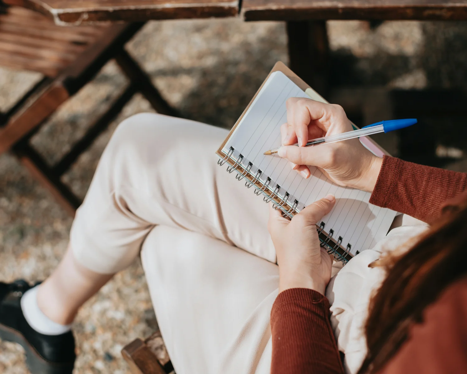 A person sitting outdoors, writing notes in a spiral notebook